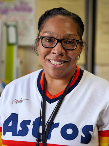 Smiling woman standing in a school hallway, wearing a white Astros jersey with lockers in the background.