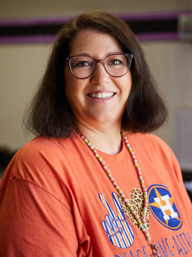 Smiling woman standing in a school hallway, wearing a orange “Astros” jersey, with student artwork displayed on lockers behind her.