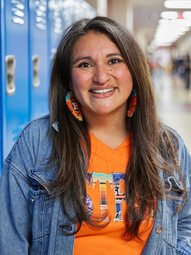 Smiling woman standing in a school hallway, wearing an orange Astro's t-shirt with a denim jacket and fun earrings.