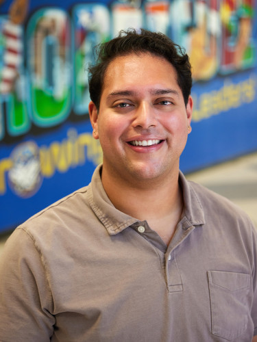 Smiling man standing in a school hallway, wearing a brown polo shirt with school artwork in the background.