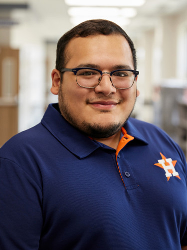 Smiling man standing in a school classroom, wearing a navy blue Astros polo with orange accents and glasses.