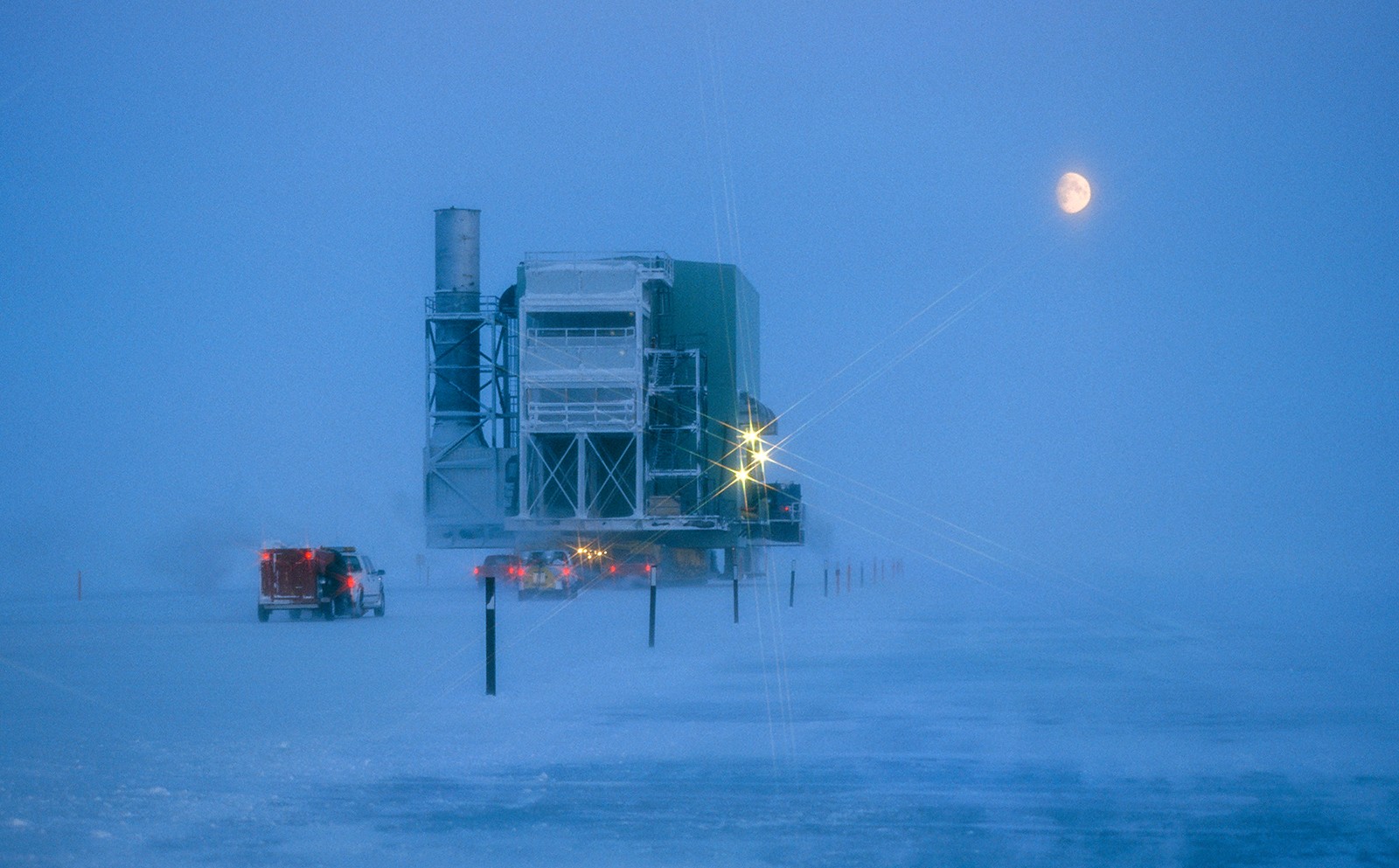 Large structure and vehicles traveling on ice road in low visibility snowy conditions, moon visible in sky