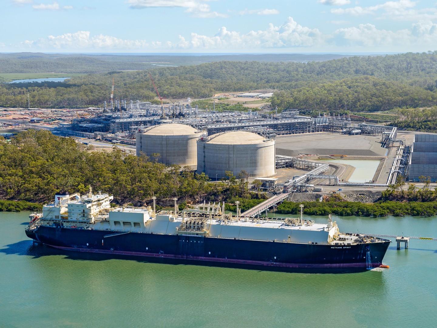 LNG ship at dock in foreground with tanks and facility in background surrounded by trees.