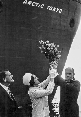 Black and white photo with three people in foreground. A man and woman are lifting a bouquet of flowers. In the background is the ship's bow with the name Arctic Tokyo.
