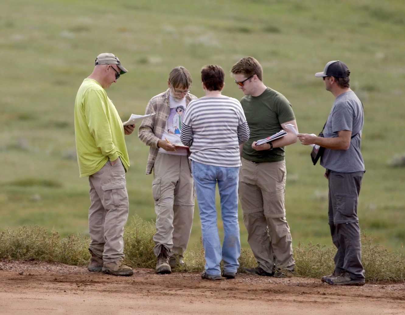 Group of five people conversing, standing outdoors with green field in background