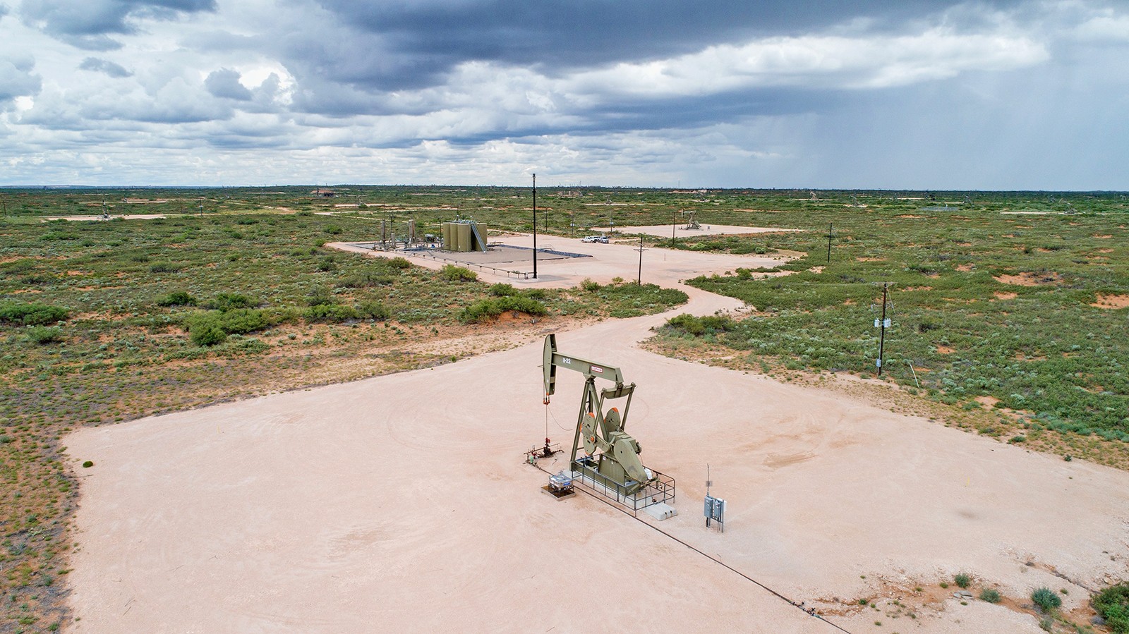 Low aerial view of pad with pumpjack connected to pad with battery tanks further away and green scrubby landscape surrounding