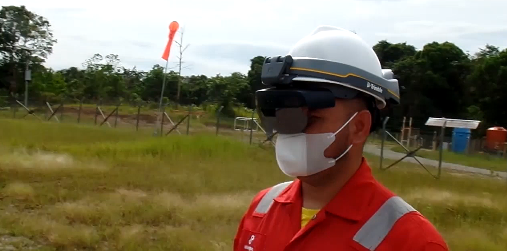 worker in hard hat, goggles and mask, outdoors with greenery and fencing in background