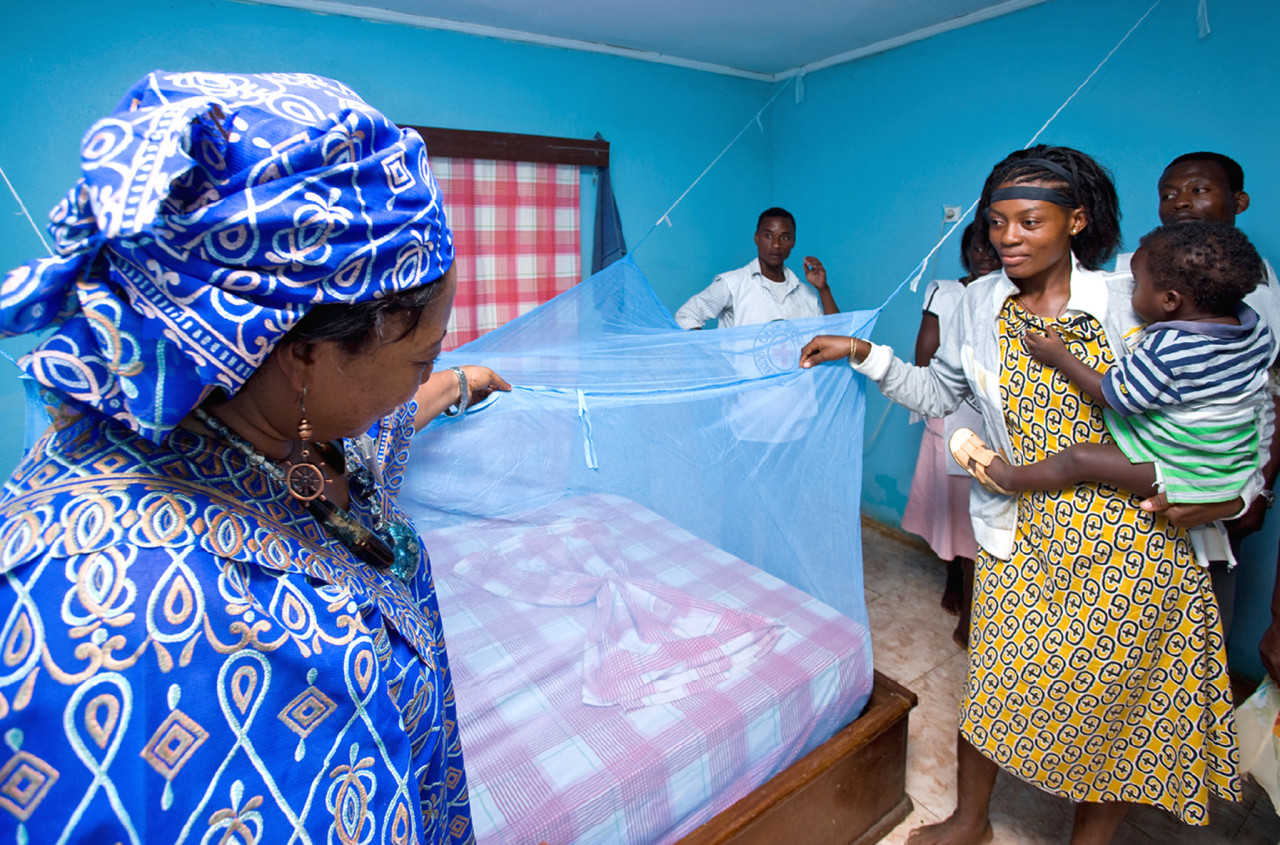 Two women examine netting over a bed. One is holding a child and three other people are in the background