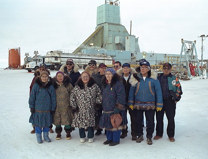 Group of thirteen people gathered on snow with rig in background