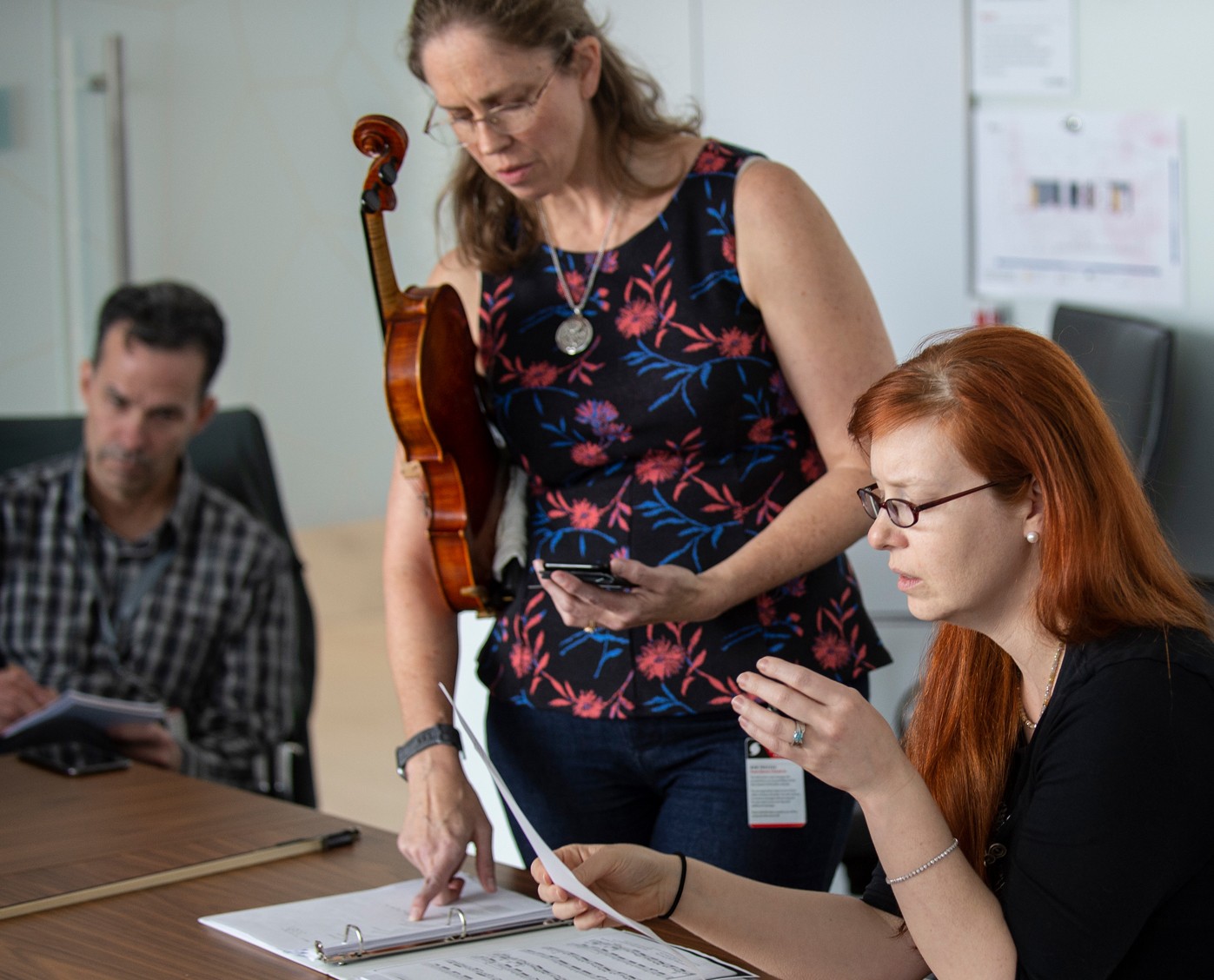 Stephanie standing holding violin, pointing at sheet music, while Bret and Kat are seated at conference table.